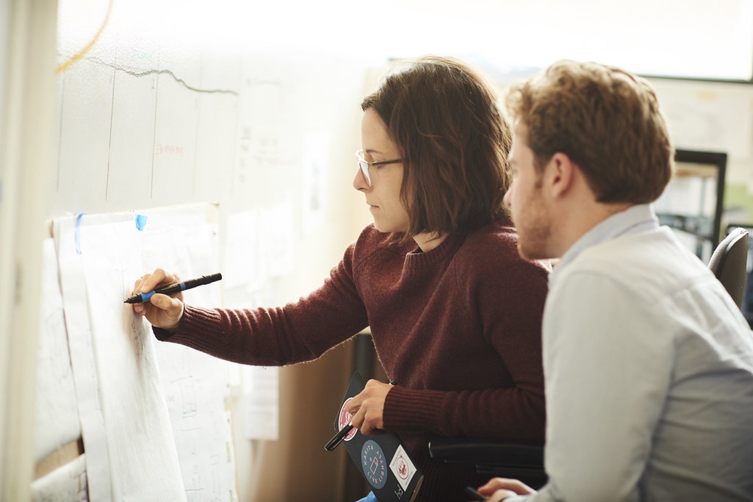 Två yrkesverksamma personer som samarbetar vid en whiteboard eller planeringstavla med diagram eller grafer. En person skriver eller ritar medan den andra observerar noggrant, vilket visar på lagarbete och kollaborativ problemlösning – viktiga delar av en innovativ arbetskultur.