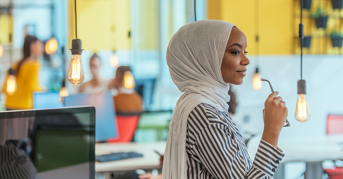 En professionell kvinna i hijab står fundersamt på en modern, ljus arbetsplats med stilfull takbelysning. I bakgrunden syns ett öppet kontorslandskap med färgglada möbler och andra medarbetare, vilket representerar mångfald i en innovativ arbetsmiljö med samtida designelement.